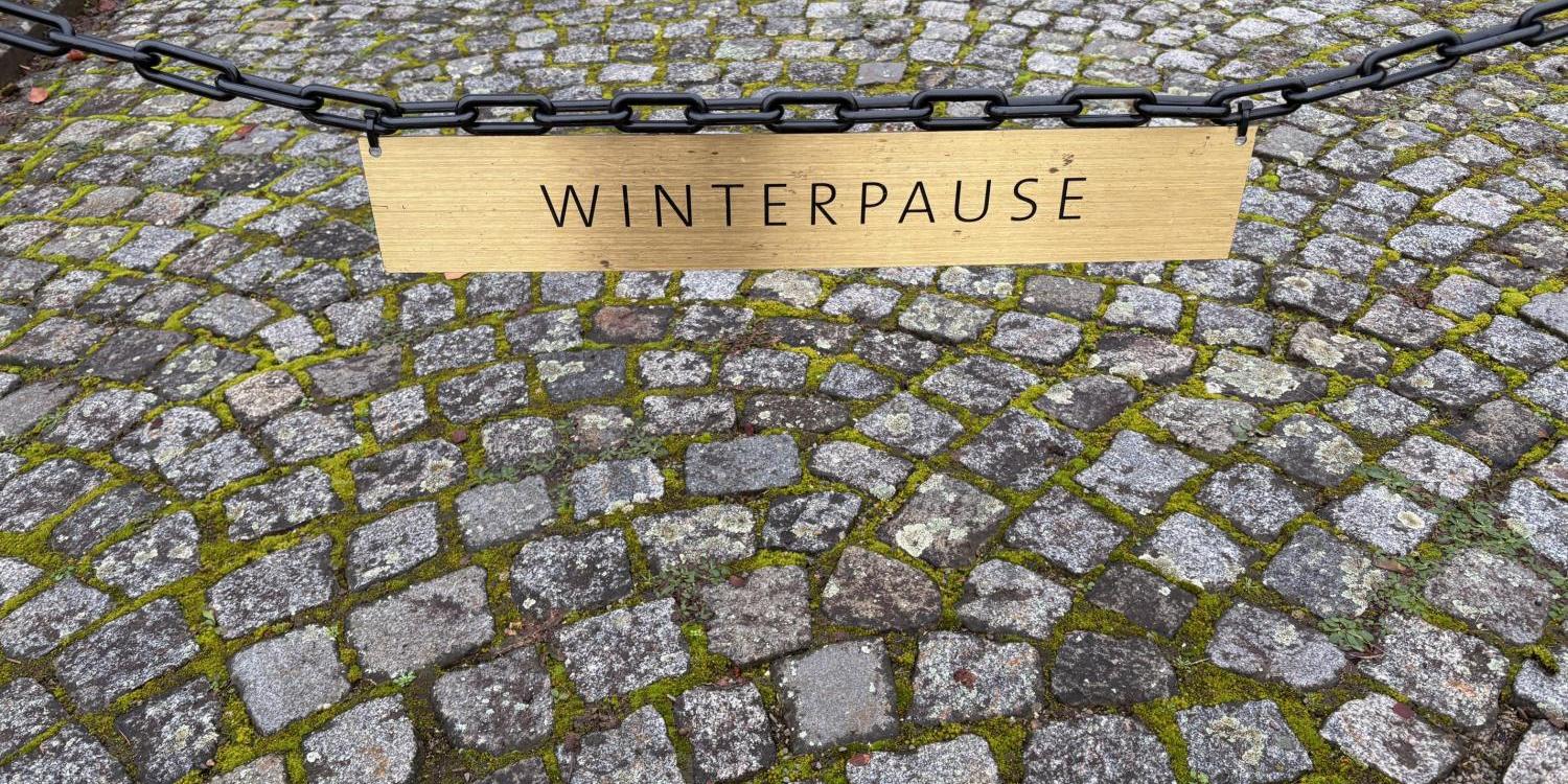 Das Bild zeigt eine Kette, die einen Weg aus Kopfsteinpflaster absperrt. An der Kette hängt ein Holzschild mit der Aufschrift „WINTERPAUSE“ in schwarzen Buchstaben.