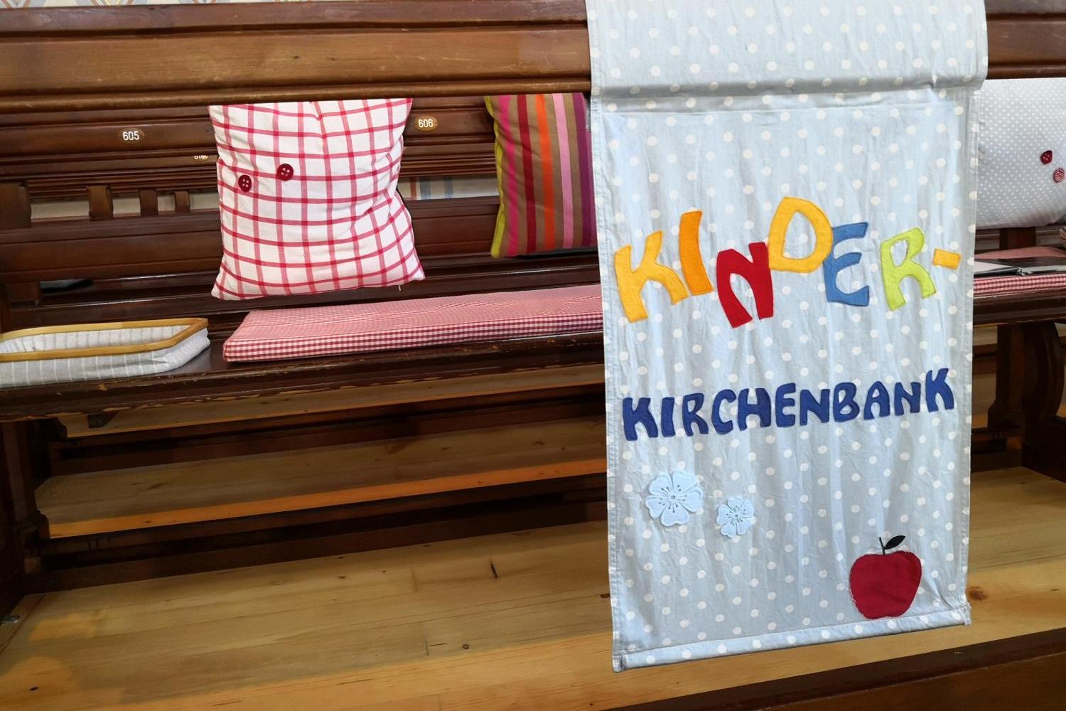 Auf einer Kirchenbank liegen Kissen, und davor hängt ein Banner mit der Aufschrift ‚Kinderkirchenbank‘