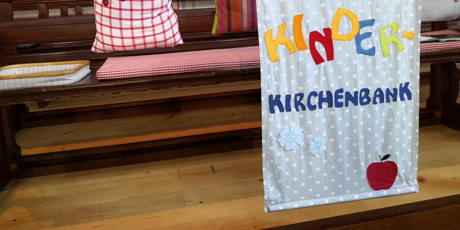 Auf einer Kirchenbank liegen Kissen, und davor hängt ein Banner mit der Aufschrift ‚Kinderkirchenbank‘