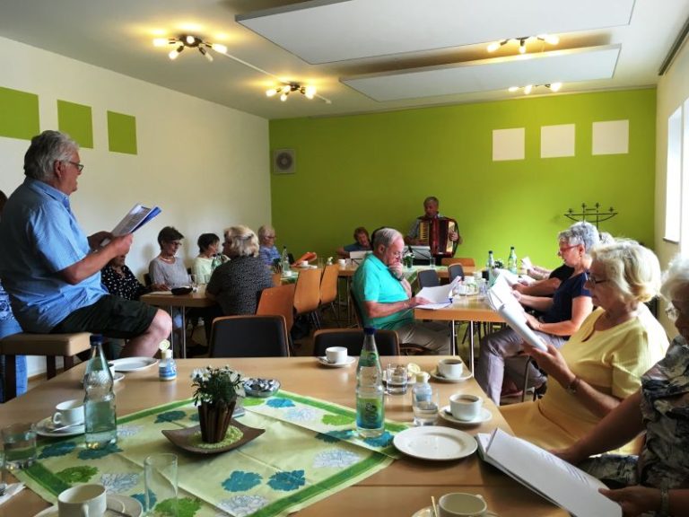 Eine Gruppe von Menschen sitzt gemeinsam in einem gemütlich gedeckten Raum, trinkt Kaffee und singt oder musiziert, während jemand mit einem Akkordeon spielt.