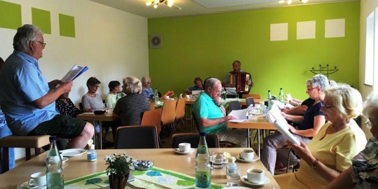 Eine Gruppe von Menschen sitzt gemeinsam in einem gemütlich gedeckten Raum, trinkt Kaffee und singt oder musiziert, während jemand mit einem Akkordeon spielt.