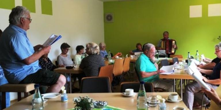Eine Gruppe von Menschen sitzt gemeinsam in einem gemütlich gedeckten Raum, trinkt Kaffee und singt oder musiziert, während jemand mit einem Akkordeon spielt.