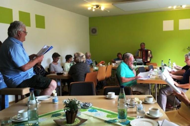 Eine Gruppe von Menschen sitzt gemeinsam in einem gemütlich gedeckten Raum, trinkt Kaffee und singt oder musiziert, während jemand mit einem Akkordeon spielt.
