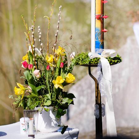 Eine große, bunt verzierte Osterkerze steht neben einem Strauß aus gelben und rosa Frühlingsblumen.