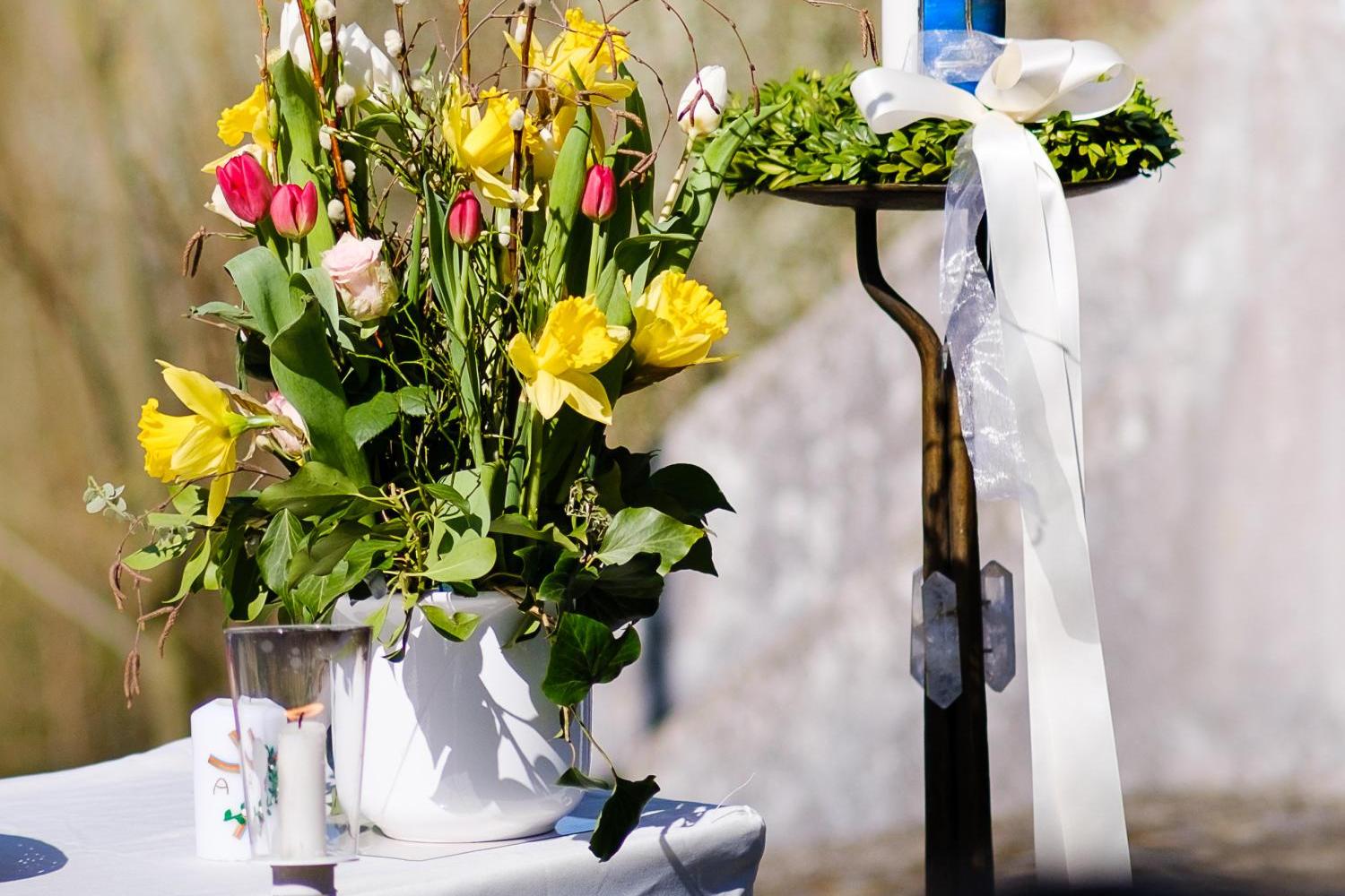 Eine große, bunt verzierte Osterkerze steht neben einem Strauß aus gelben und rosa Frühlingsblumen.