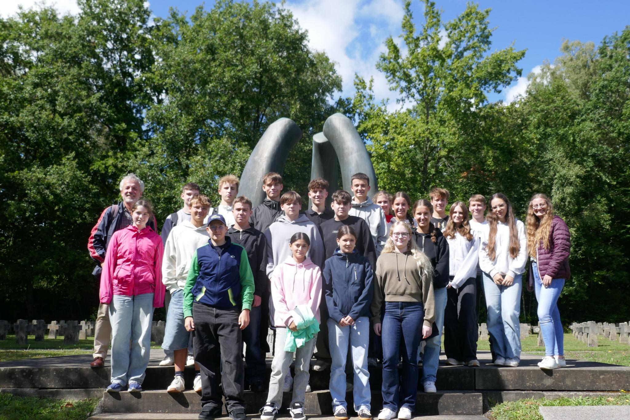 Das Foto zeigt eine Gruppe junger Menschen in Freizeitkleidung, die draußen vor einer großen, abstrakten Metallskulptur stehen – im Hintergrund sind grüne Bäume und ein blauer Himmel mit Wolken zu sehen. (c) Thomas Kupczik Das Foto zeigt eine Gruppe junger Menschen in Freizeitkleidung, die draußen vor einer großen, abstrakten Metallskulptur stehen – im Hintergrund sind grüne Bäume und ein blauer Himmel mit Wolken zu sehen.