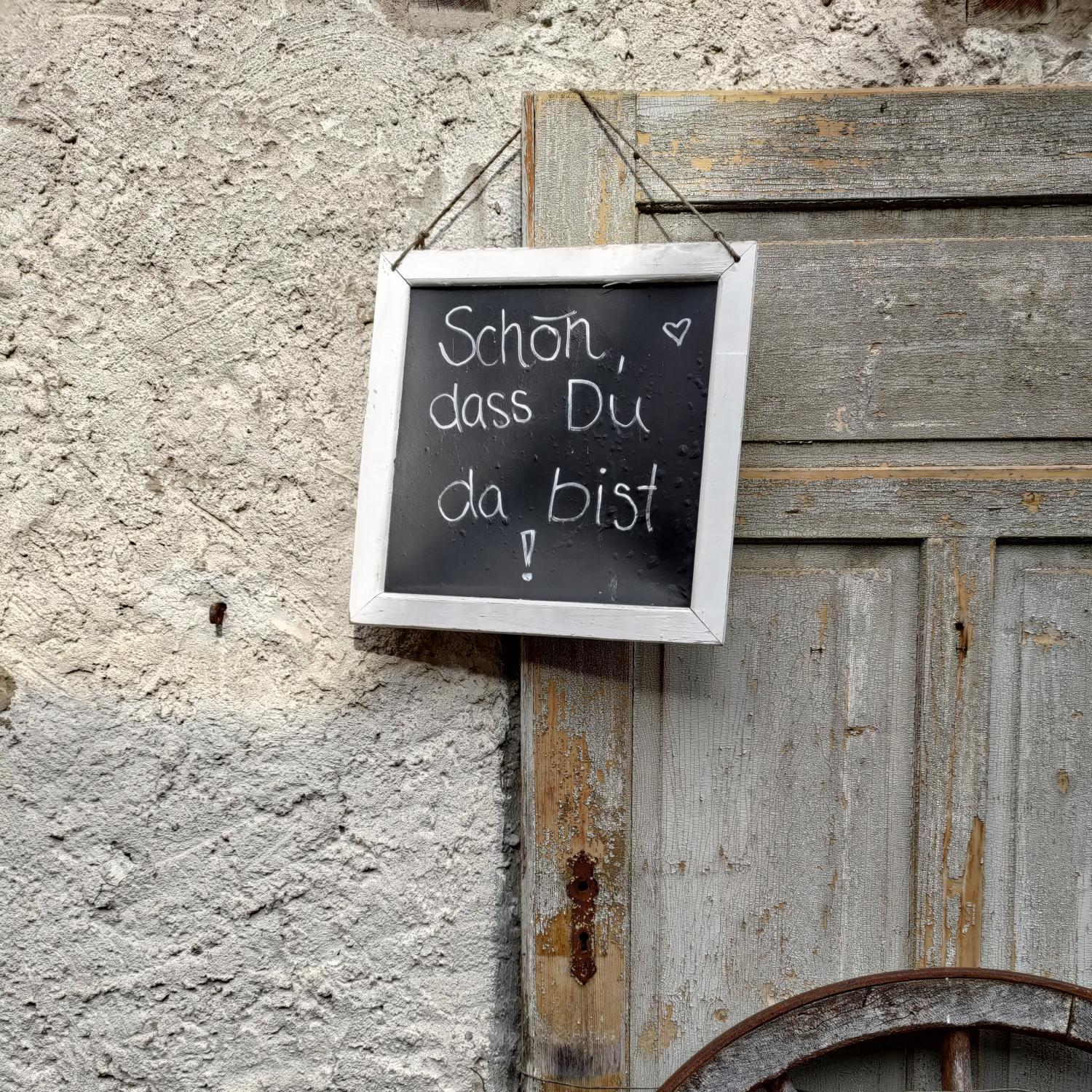 Ein Schild mit der Aufschrift „Schön, dass Du da bist!“ hängt an einer alten Holztür, daneben ein Wagenrad.