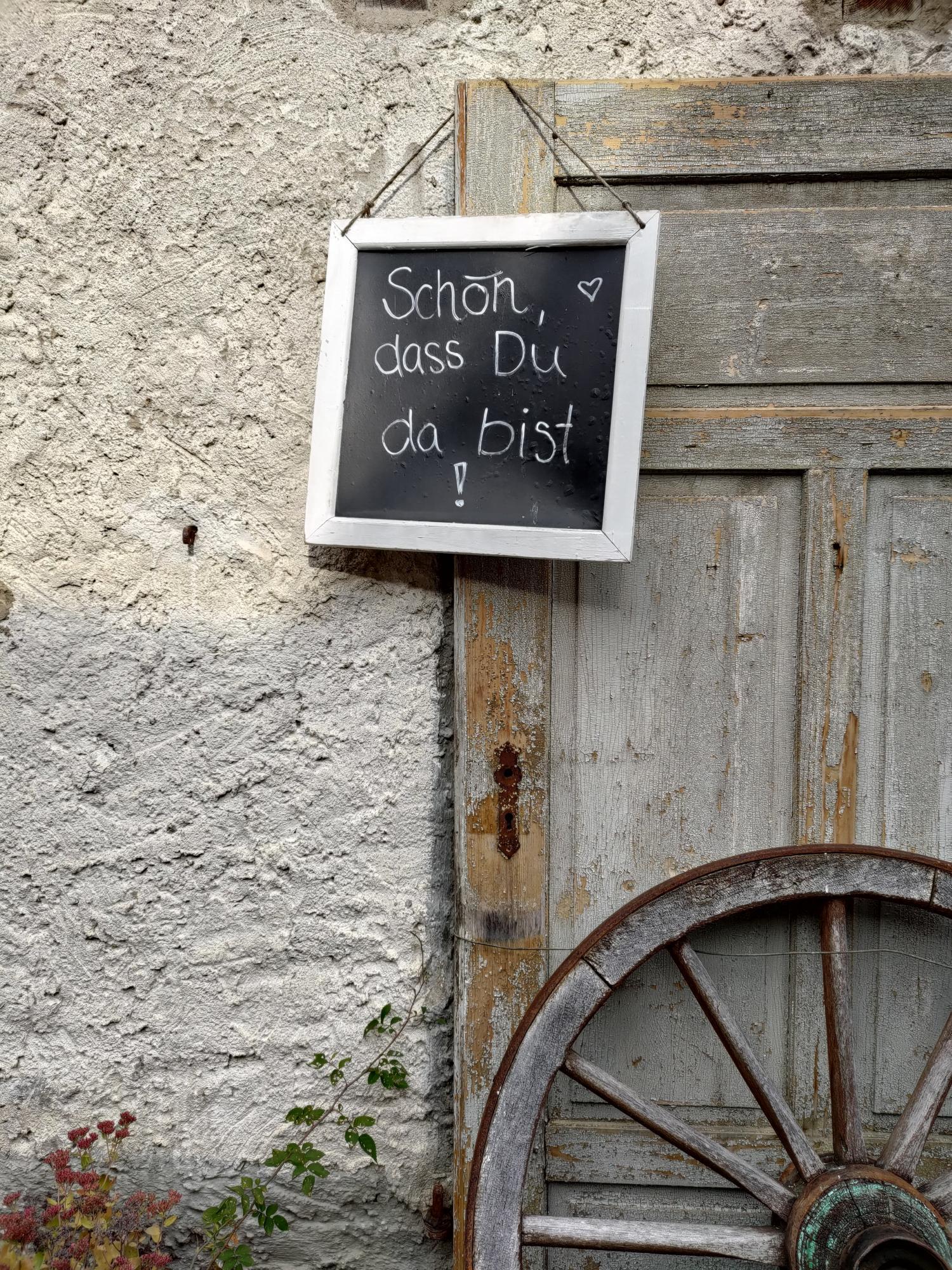 Ein Schild mit der Aufschrift „Schön, dass Du da bist!“ hängt an einer alten Holztür, daneben ein Wagenrad.
