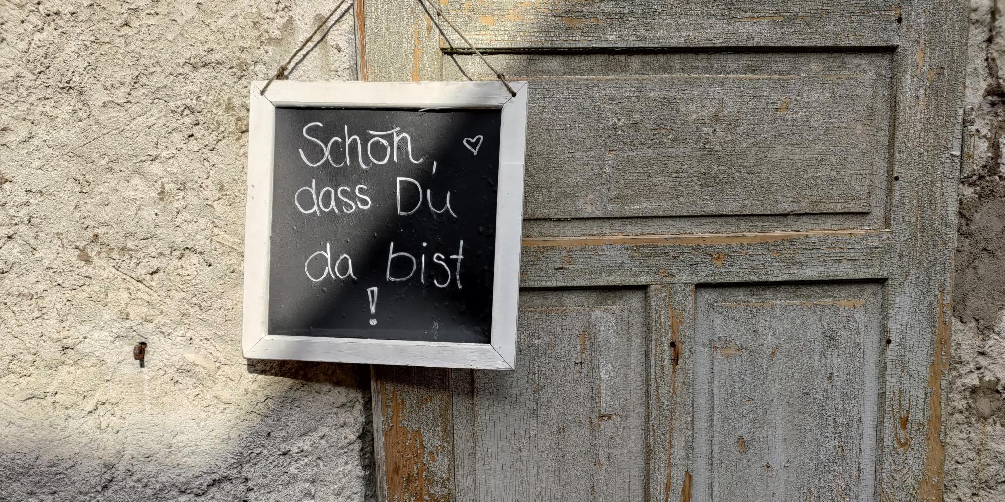 Ein kleines Willkommensschild hängt vor einer alten Holztür, daneben ein altes Wagenrad.