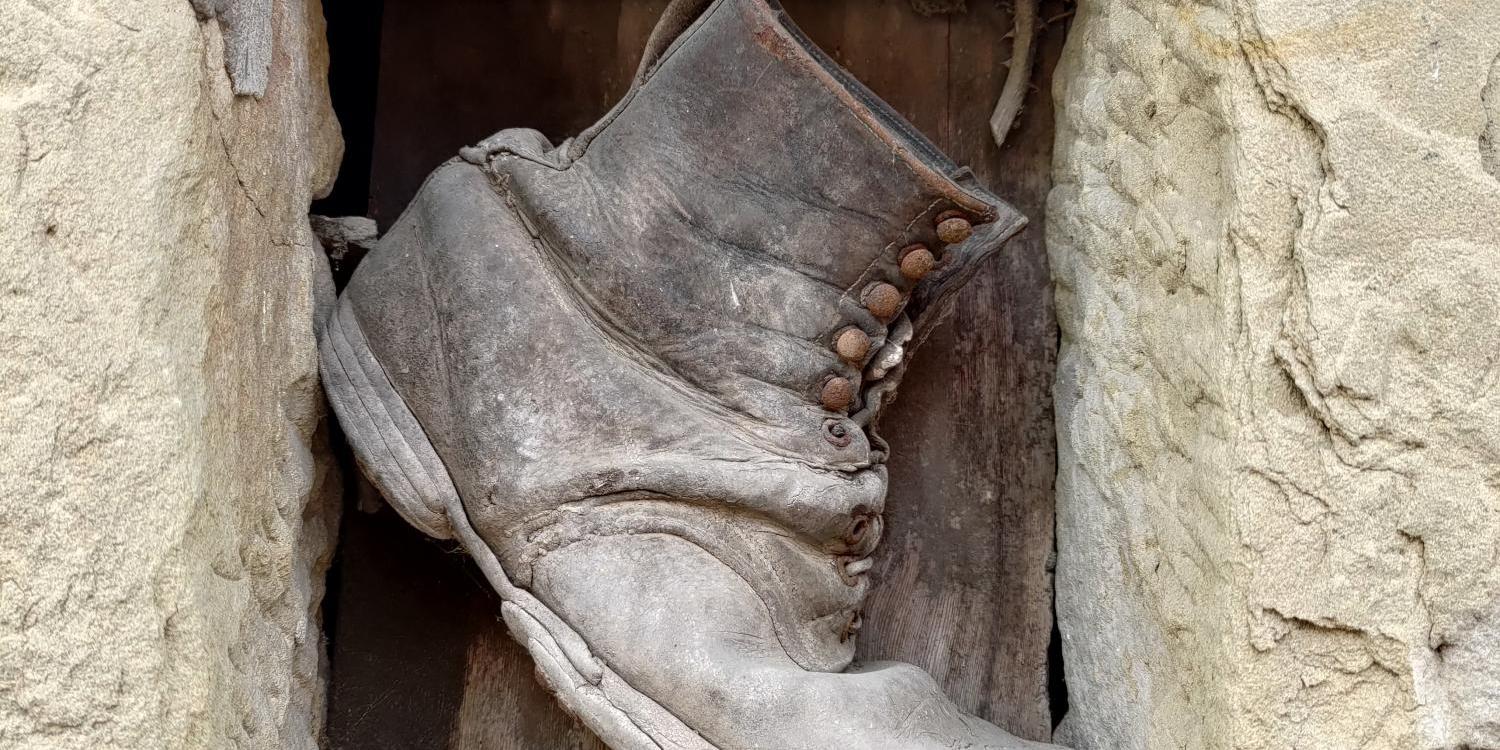 Das Foto zeigt einen alten, stark abgenutzten Wanderstiefel, der zwischen groben Steinblöcken und Holz in einer Mauer eingeklemmt ist.