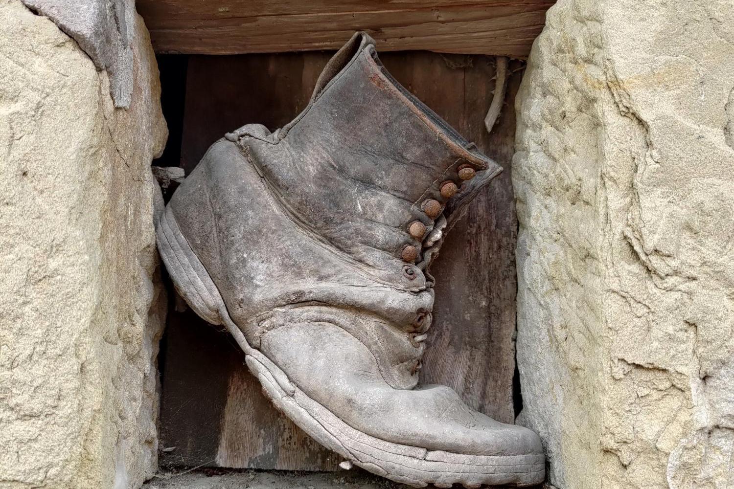 Das Foto zeigt einen alten, stark abgenutzten Wanderstiefel, der zwischen groben Steinblöcken und Holz in einer Mauer eingeklemmt ist.