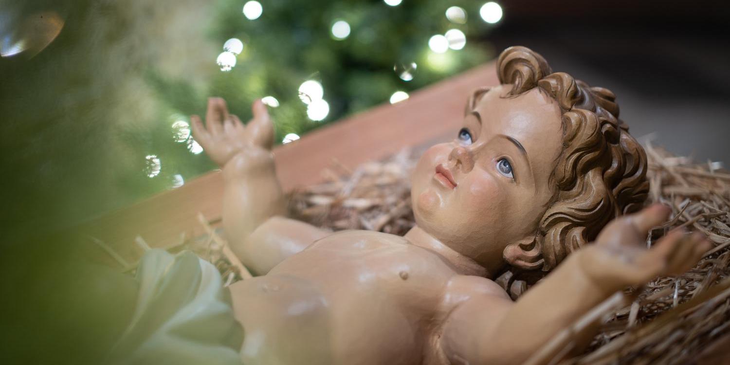 Das Bild zeigt eine Krippenszene mit dem Jesuskind in einer einfachen Futterkrippe im Vordergrund, während im Hintergrund ein festlich geschmückter und leuchtender Weihnachtsbaum steht. (c) Markus Weinländer In: Pfarrbriefservice.de Das Bild zeigt eine Krippenszene mit dem Jesuskind in einer einfachen Futterkrippe im Vordergrund, während im Hintergrund ein festlich geschmückter und leuchtender Weihnachtsbaum steht.