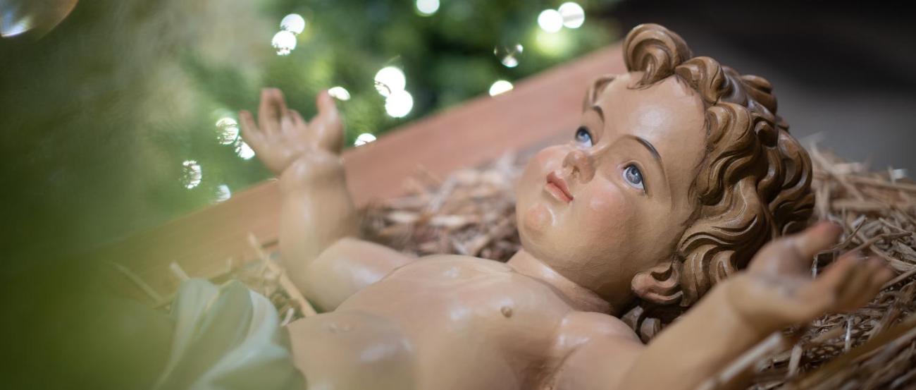 Das Bild zeigt eine Krippenszene mit dem Jesuskind in einer einfachen Futterkrippe im Vordergrund, während im Hintergrund ein festlich geschmückter und leuchtender Weihnachtsbaum steht.