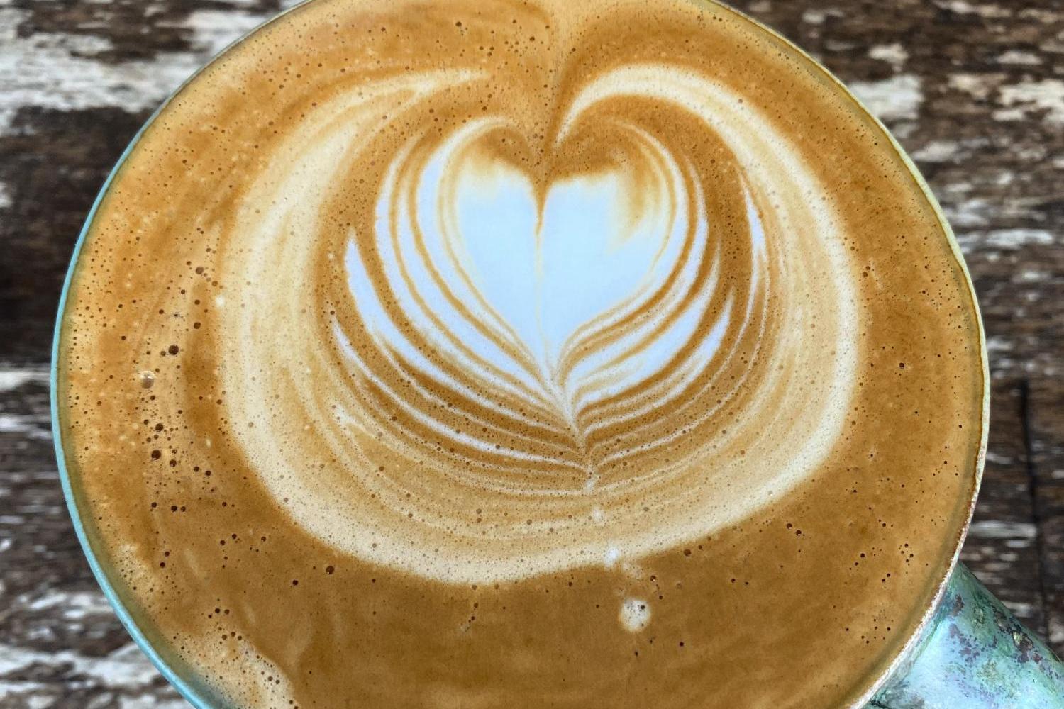 Das Foto zeigt eine Tasse Kaffee mit kunstvoller Latte-Art in Form eines Herzens auf einer rustikalen Holzoberfläche.