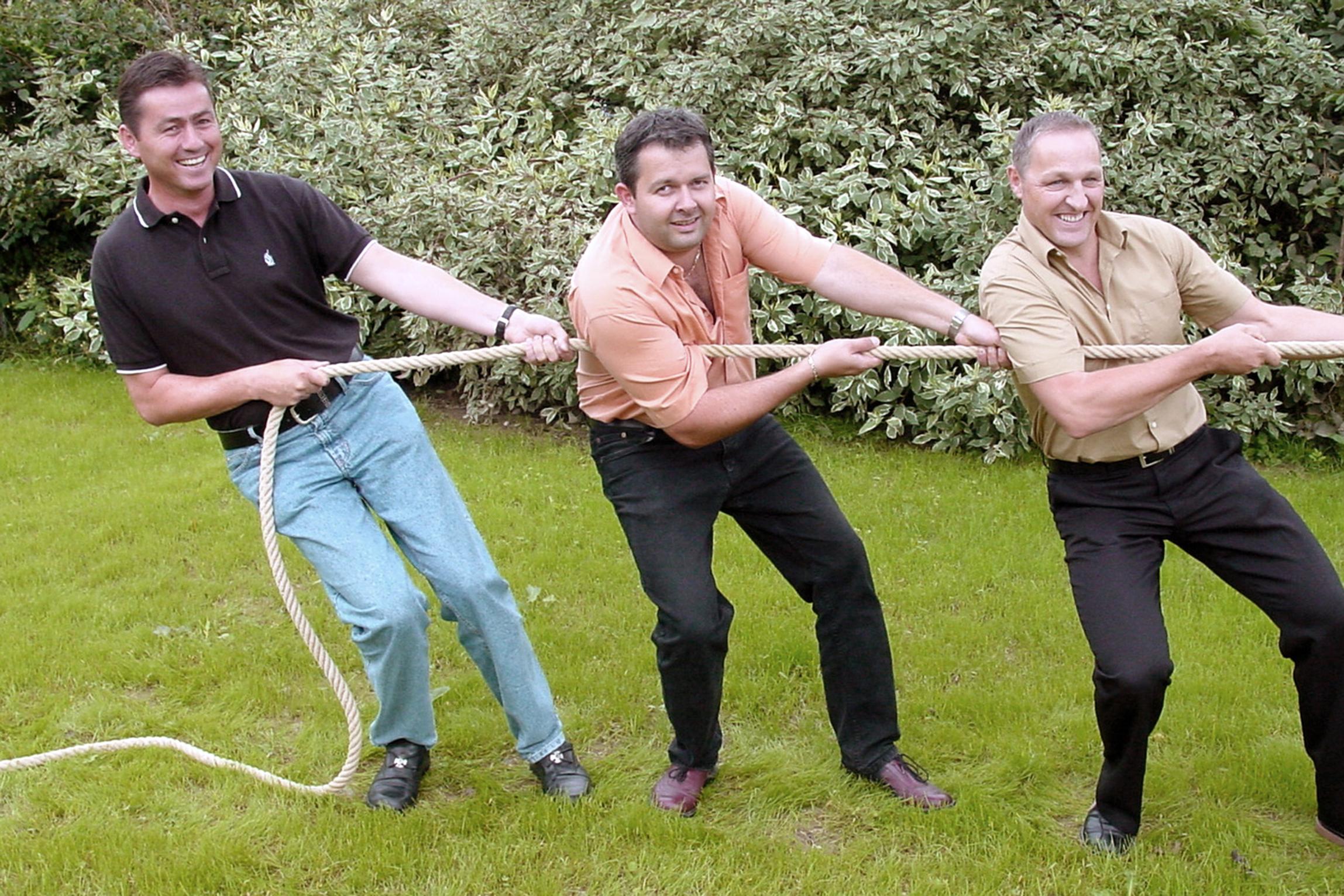 Das Bild zeigt drei Männer, die auf einer Wiese stehen und gemeinsam an einem Seil ziehen.