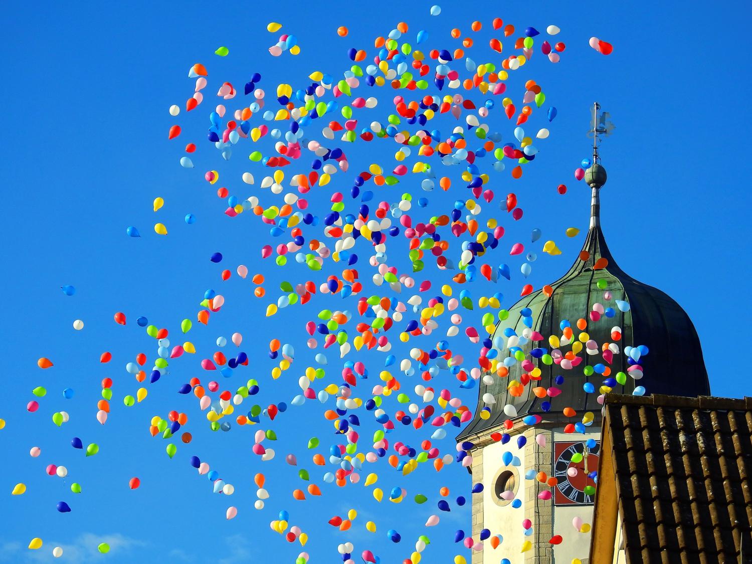 Viele bunte Luftballons steigen in den Himmel neben einem Kirchturm mit bunt bemalter Uhr.