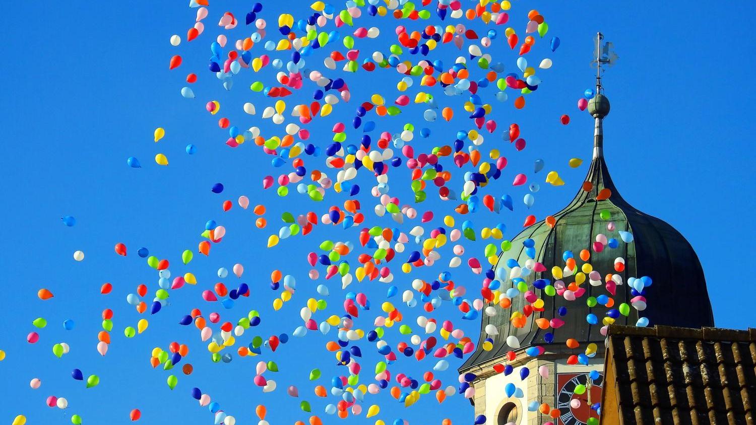 Viele bunte Luftballons steigen in den Himmel neben einem Kirchturm mit bunt bemalter Uhr.