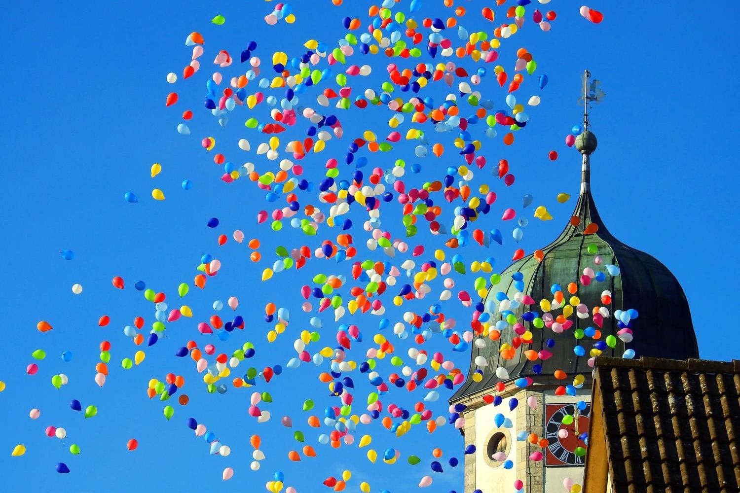 Viele bunte Luftballons steigen in den Himmel neben einem Kirchturm mit bunt bemalter Uhr.