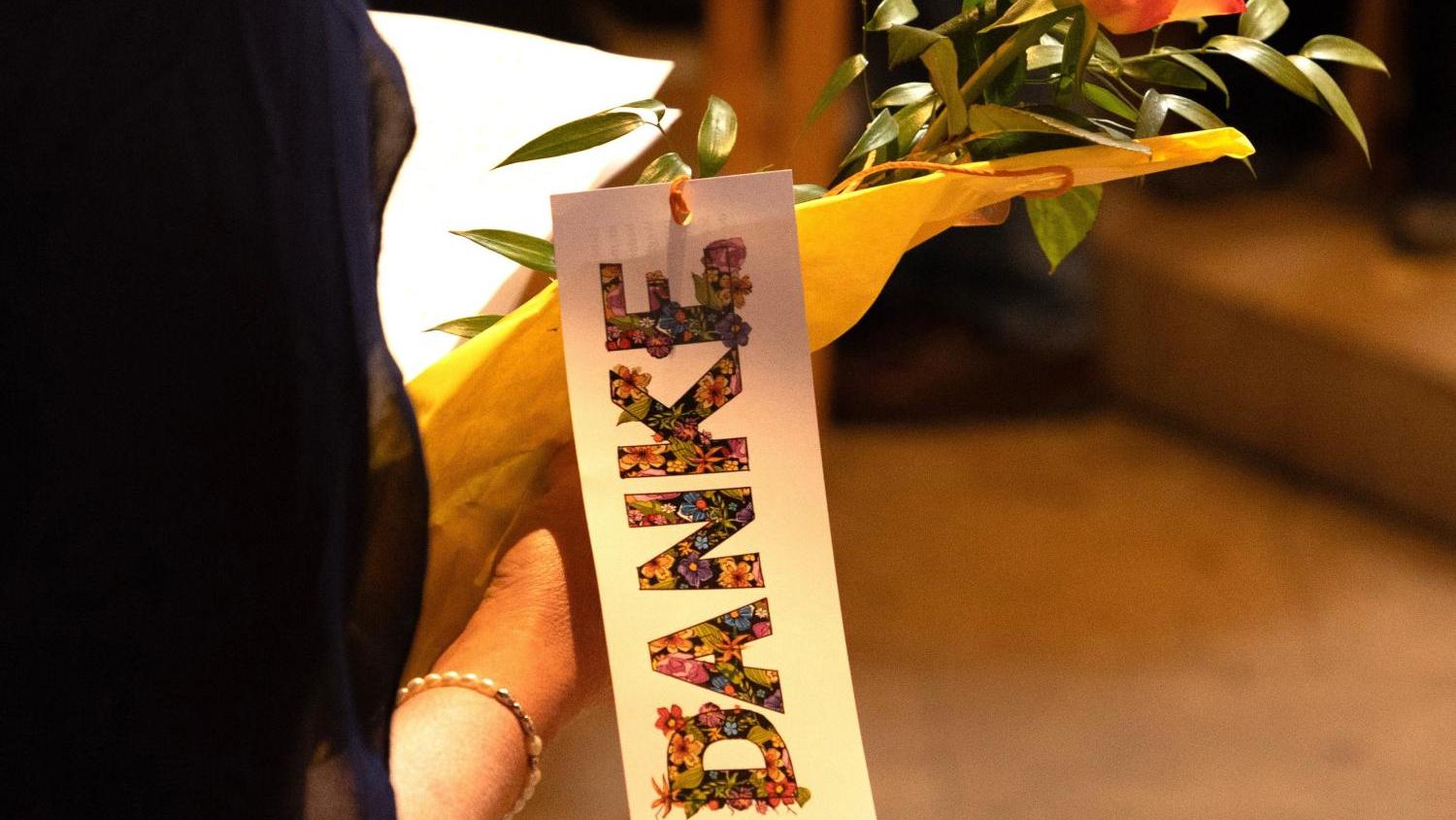 Eine Person hält eine orangefarbene Rose, eingewickelt in gelbes Papier. Daran hängt ein Schild mit dem Wort „DANKE“ in bunten Blumenbuchstaben, der Hintergrund ist unscharf.