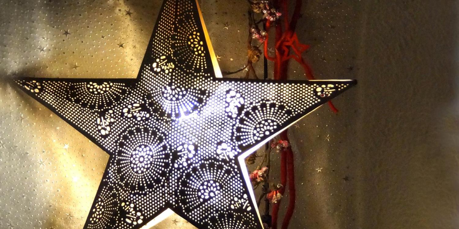 Zwei große, dunkle Metallsterne mit feinen Lochmustern leuchten warm von innen vor einem goldenen Hintergrund.