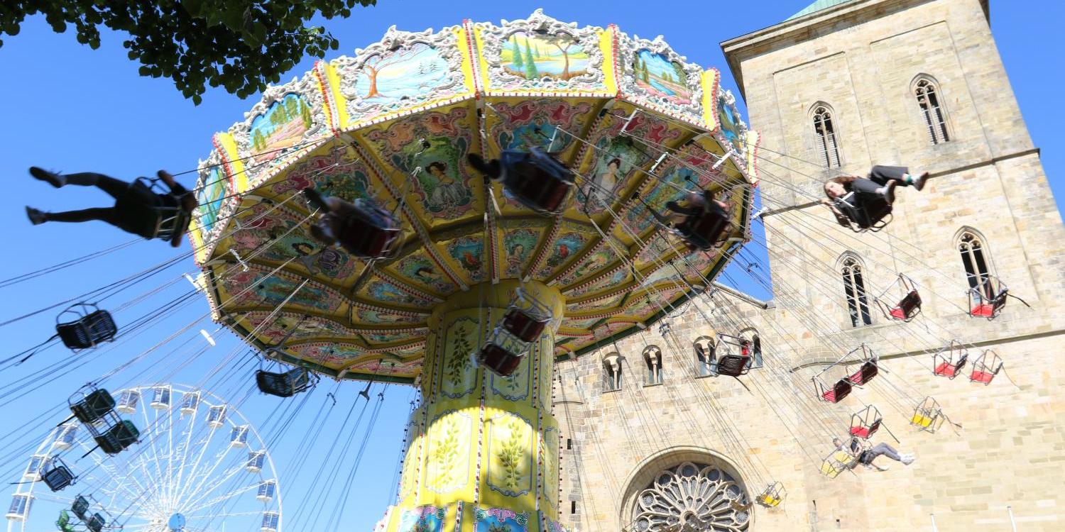 Kettenkarussell mit Menschen vor einer Kirche bei sonnigem Wetter, Riesenrad im Hintergrund.