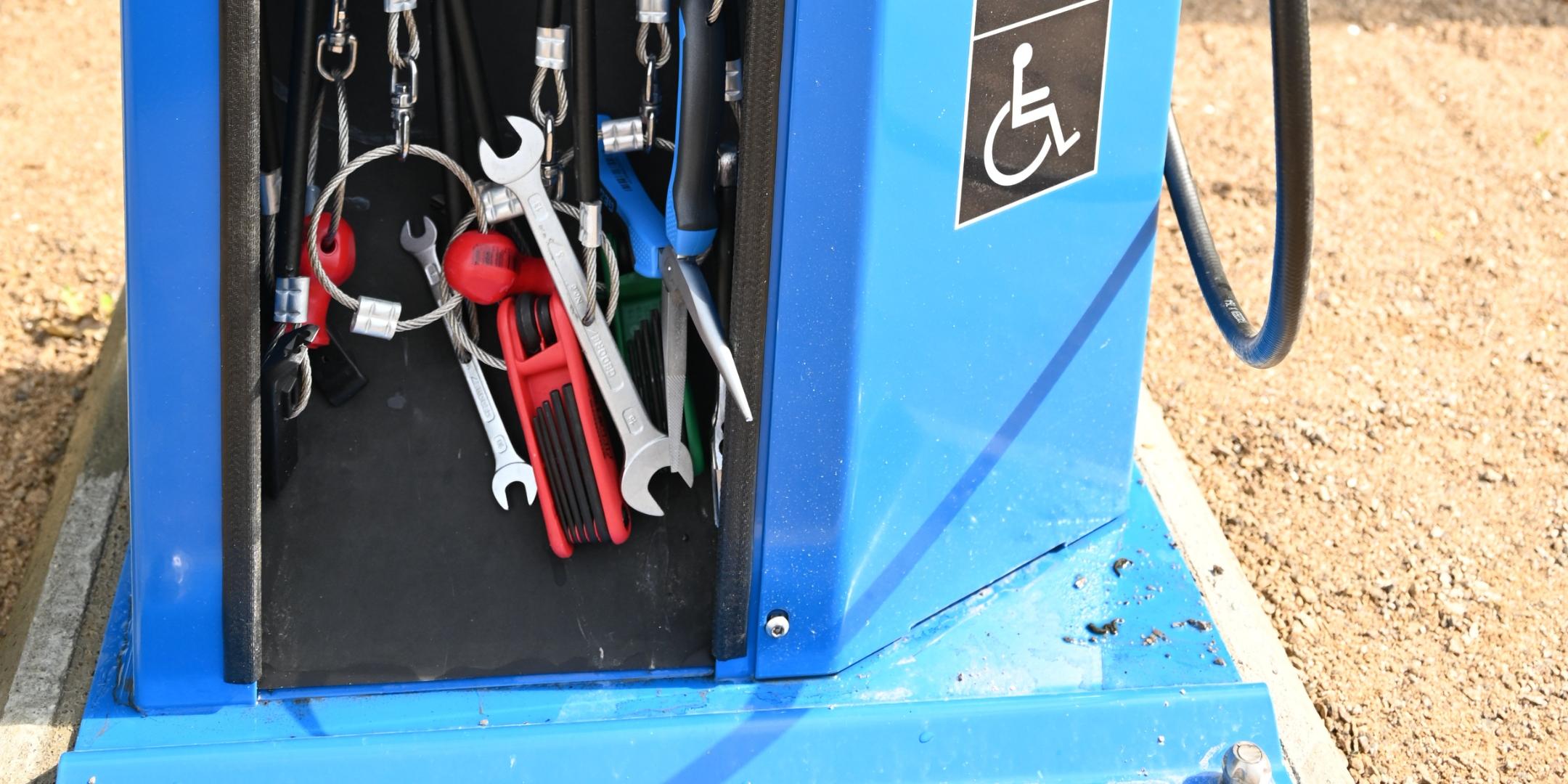 Das Foto zeigt eine blaue Fahrradreparaturstation im Freien mit befestigten Werkzeugen und einer Luftpumpe zur selbstständigen Nutzung.