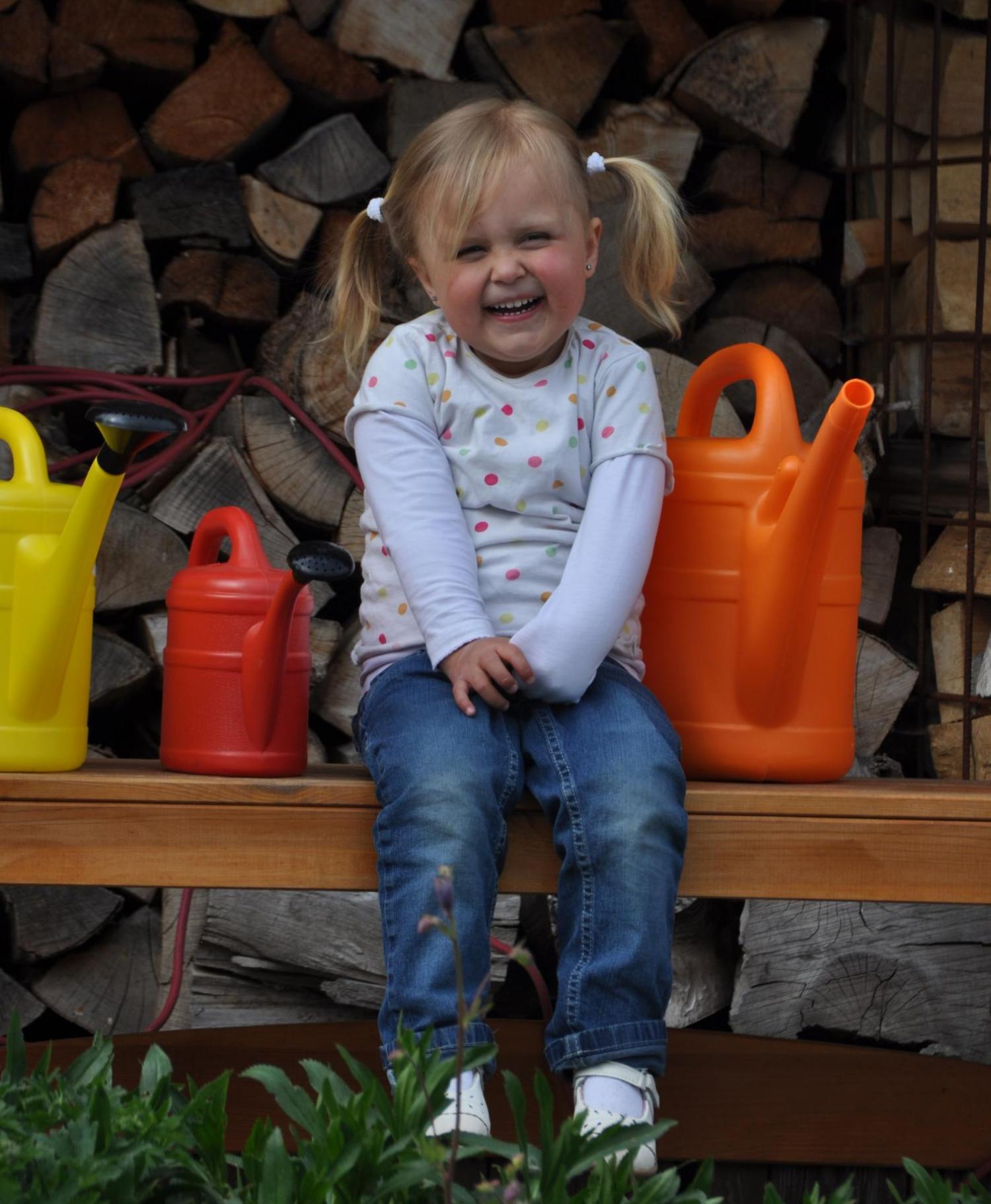 Das Foto zeigt ein kleines Kind mit Zöpfen, das auf einer Holzbank vor einem Stapel Kaminholz sitzt, umgeben von bunten Gießkannen in Gelb, Rot und Orange. (c) Michael Bogedain In: Pfarrbriefservice.de Das Foto zeigt ein kleines Kind mit Zöpfen, das auf einer Holzbank vor einem Stapel Kaminholz sitzt, umgeben von bunten Gießkannen in Gelb, Rot und Orange.