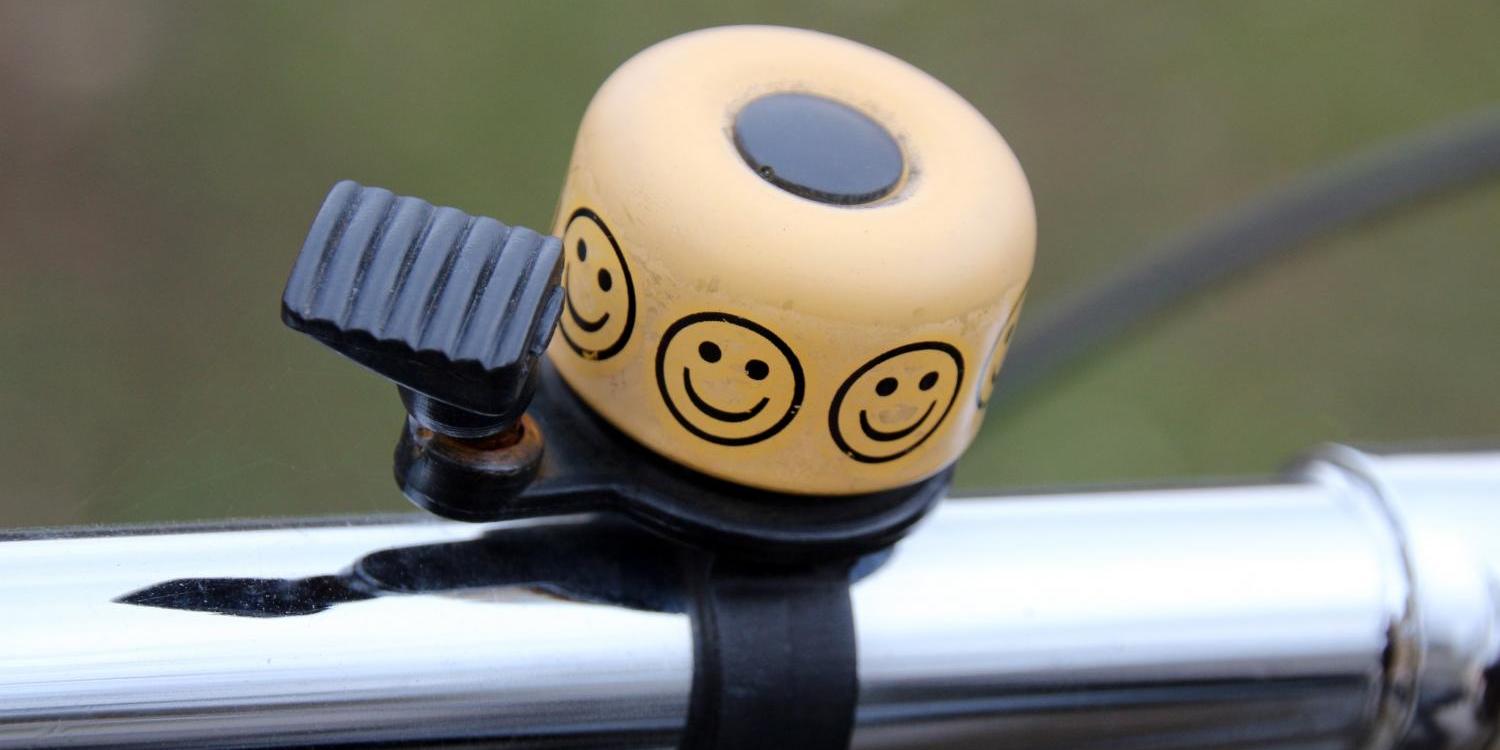 Eine gelbe Fahrradklingel mit mehreren aufgedruckten Smileys ist an einem silbernen Lenker befestigt.