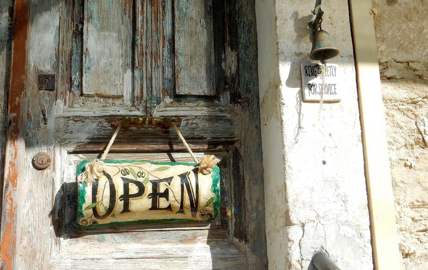 Alte Holztür mit „Open“-Schild und verblasster Hauswand im Sonnenlicht.