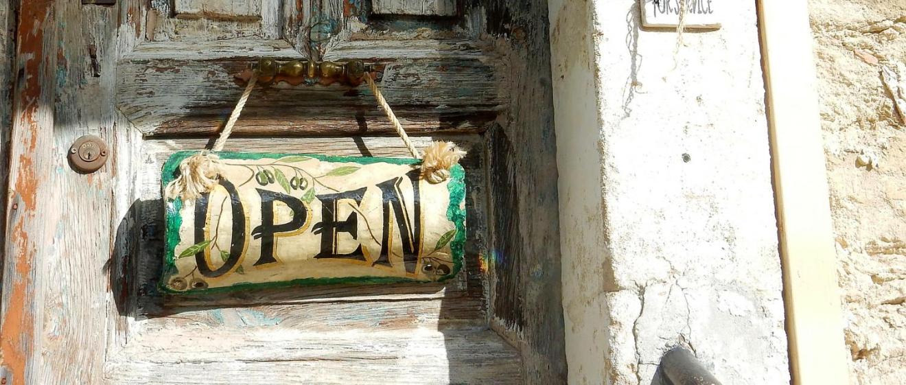 Alte Holztür mit „Open“-Schild und verblasster Hauswand im Sonnenlicht.