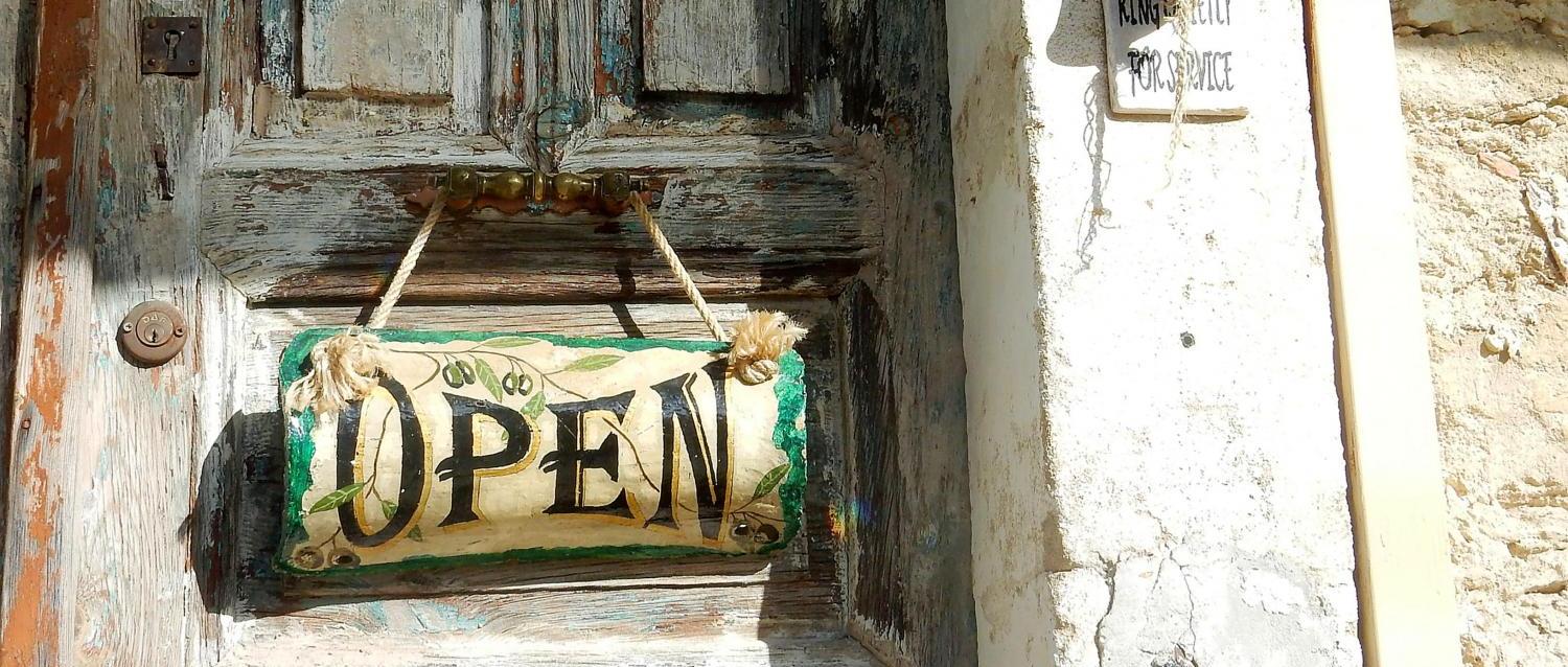 Alte Holztür mit „Open“-Schild und verblasster Hauswand im Sonnenlicht.