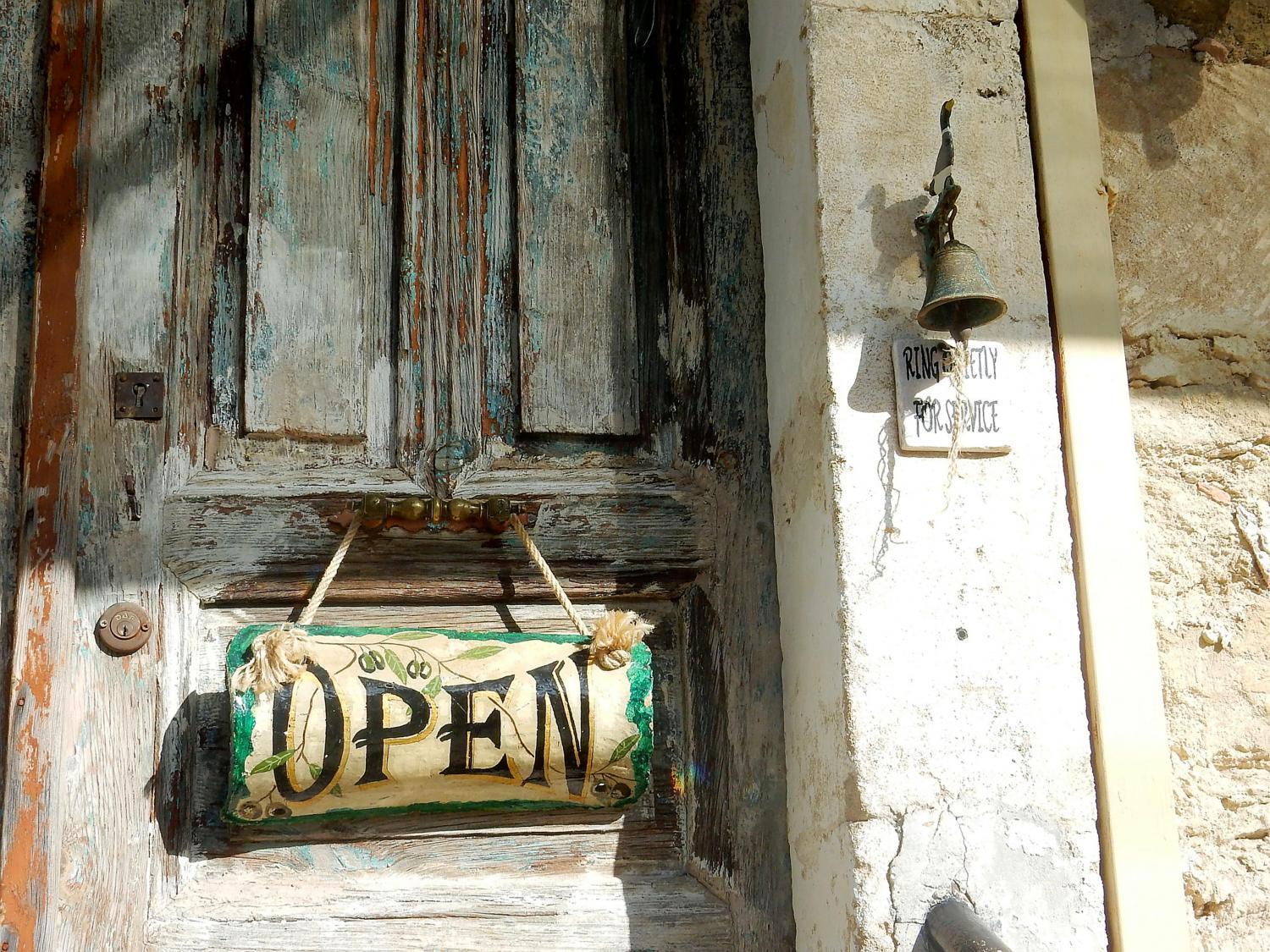Alte Holztür mit „Open“-Schild und verblasster Hauswand im Sonnenlicht.
