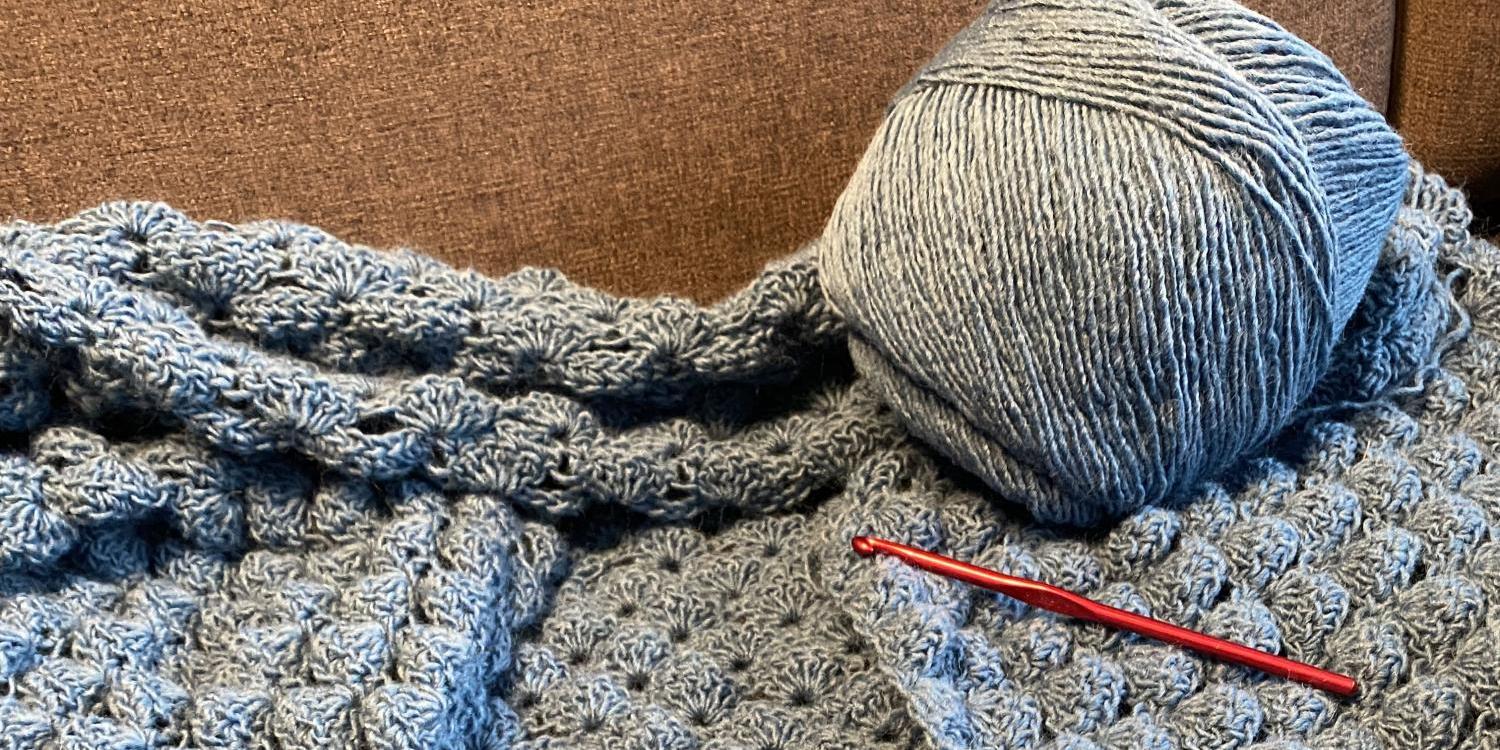 Ein graues, strukturreich gehäkeltes Stück liegt auf einem Sofa, daneben ein Wollknäuel und eine rote Häkelnadel.
