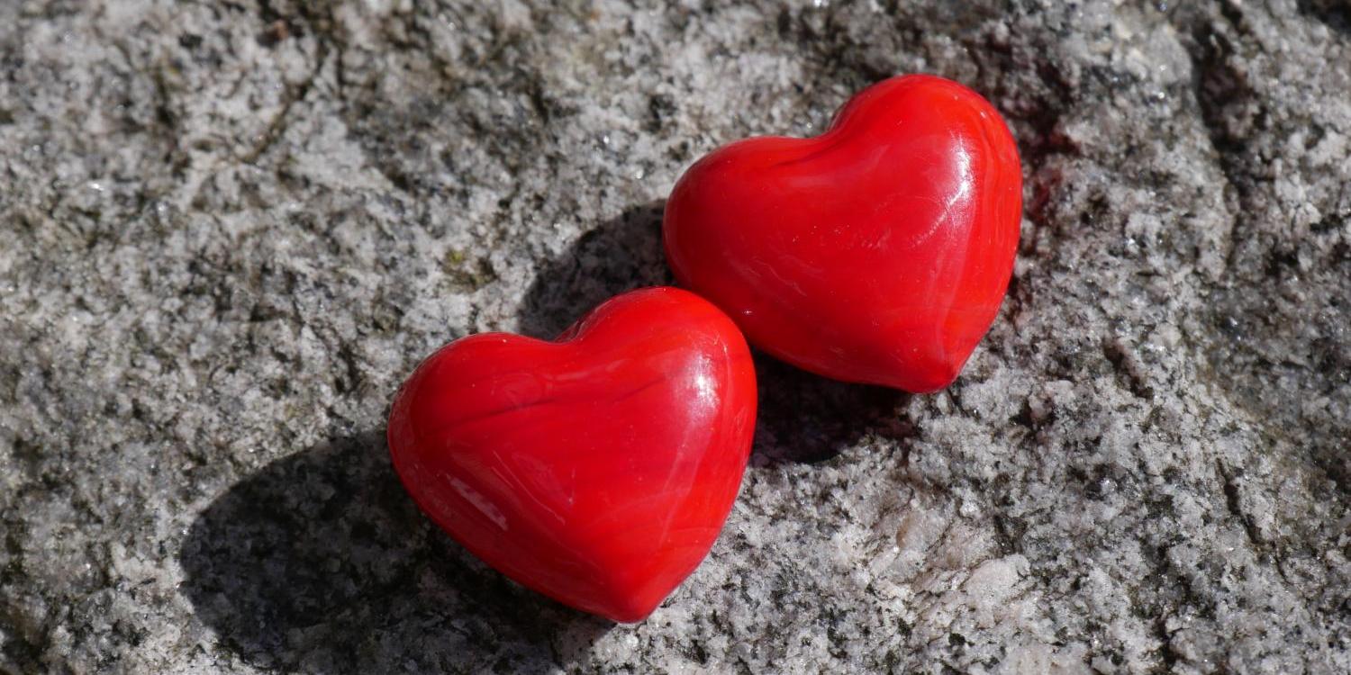 Das Foto zeigt zwei kleine, rote Herzformen, die eng nebeneinander auf einer rauen grauen Steinoberfläche liegen.