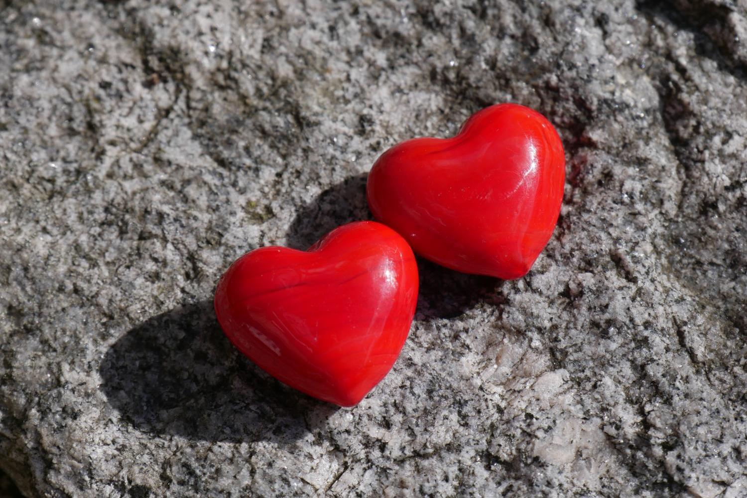 Das Foto zeigt zwei kleine, rote Herzformen, die eng nebeneinander auf einer rauen grauen Steinoberfläche liegen.
