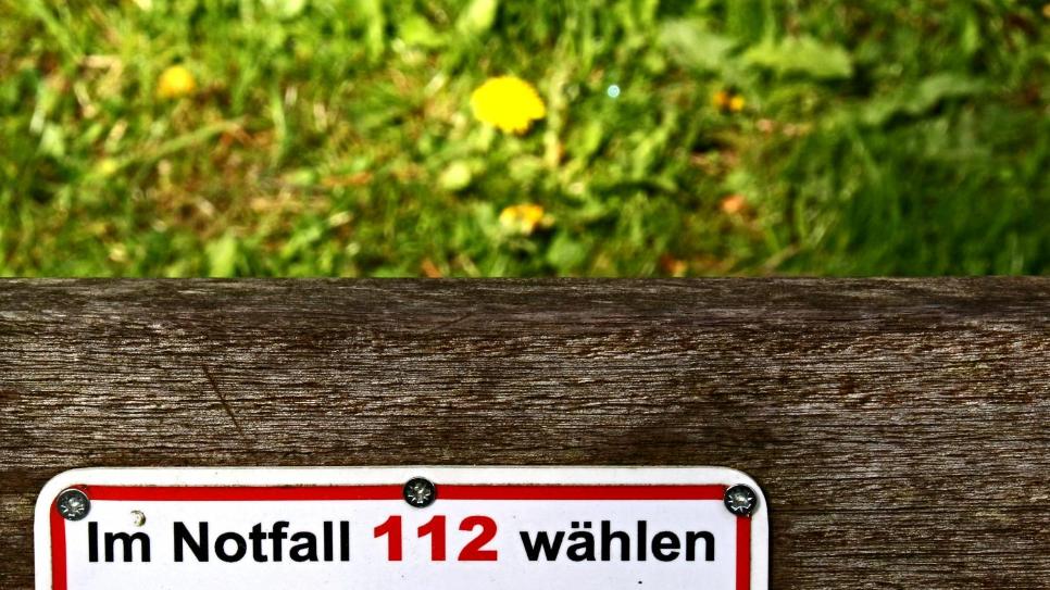 Ein Schild auf einer Holzbank trägt die Aufschrift „Im Notfall 112 wählen“.