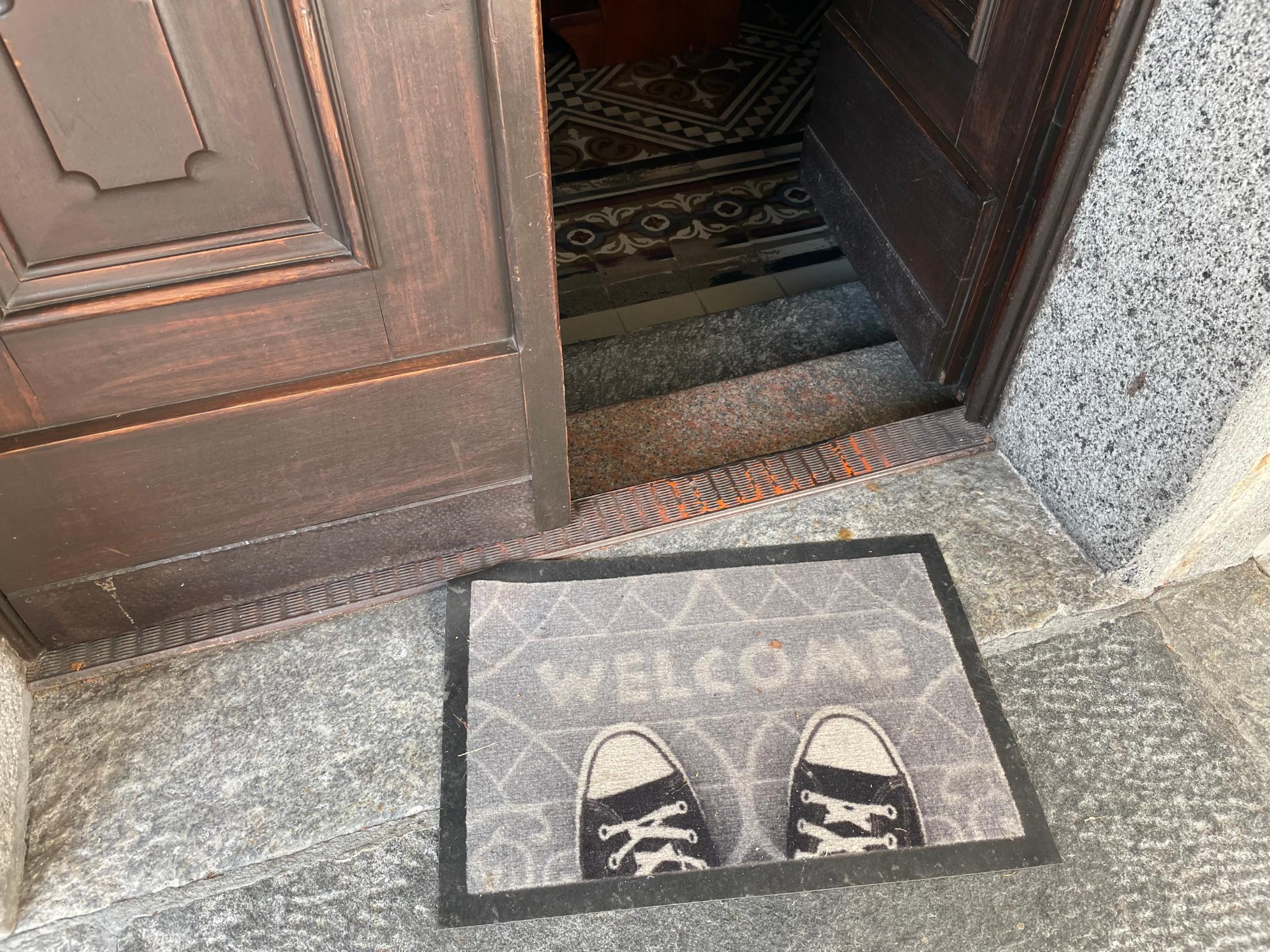 Das Foto zeigt eine geöffnete Holztür mit einer „WELCOME“-Fußmatte davor, auf der ein Paar schwarze Turnschuhe steht, dahinter führt der Eingang in ein Gebäude mit gemustertem Fliesenboden.
