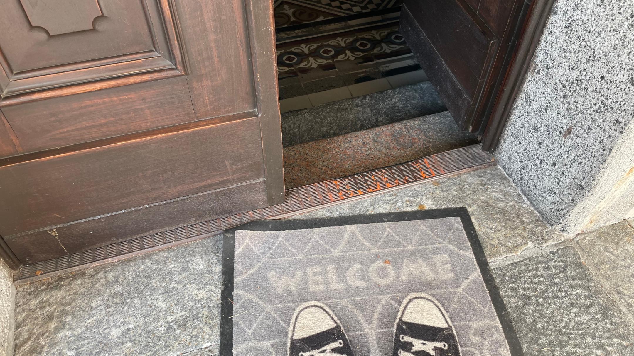 Das Foto zeigt eine geöffnete Holztür mit einer „WELCOME“-Fußmatte davor, auf der ein Paar schwarze Turnschuhe steht, dahinter führt der Eingang in ein Gebäude mit gemustertem Fliesenboden.