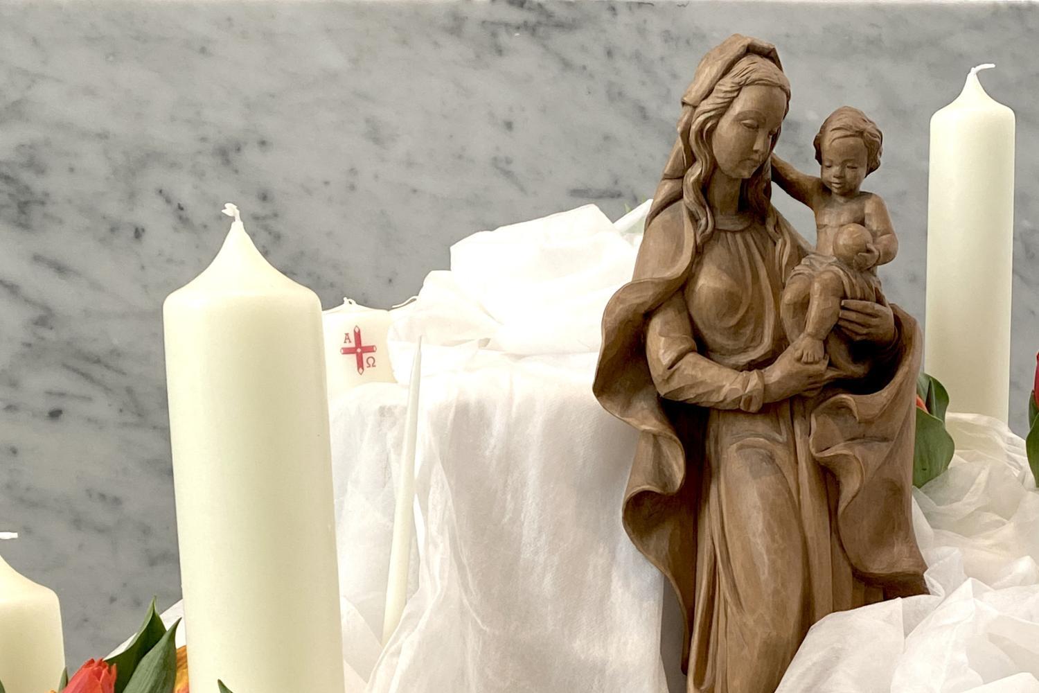 Eine hölzerne Marienstatue mit Kind steht umgeben von weißen Kerzen, Blumen und locker drapiertem weißem Stoff.