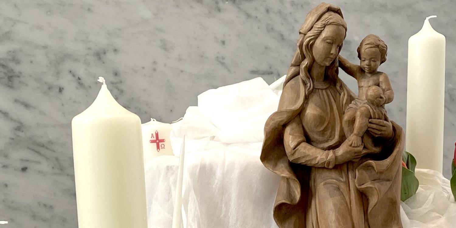 Eine hölzerne Marienstatue mit Kind steht umgeben von weißen Kerzen, Blumen und locker drapiertem weißem Stoff.