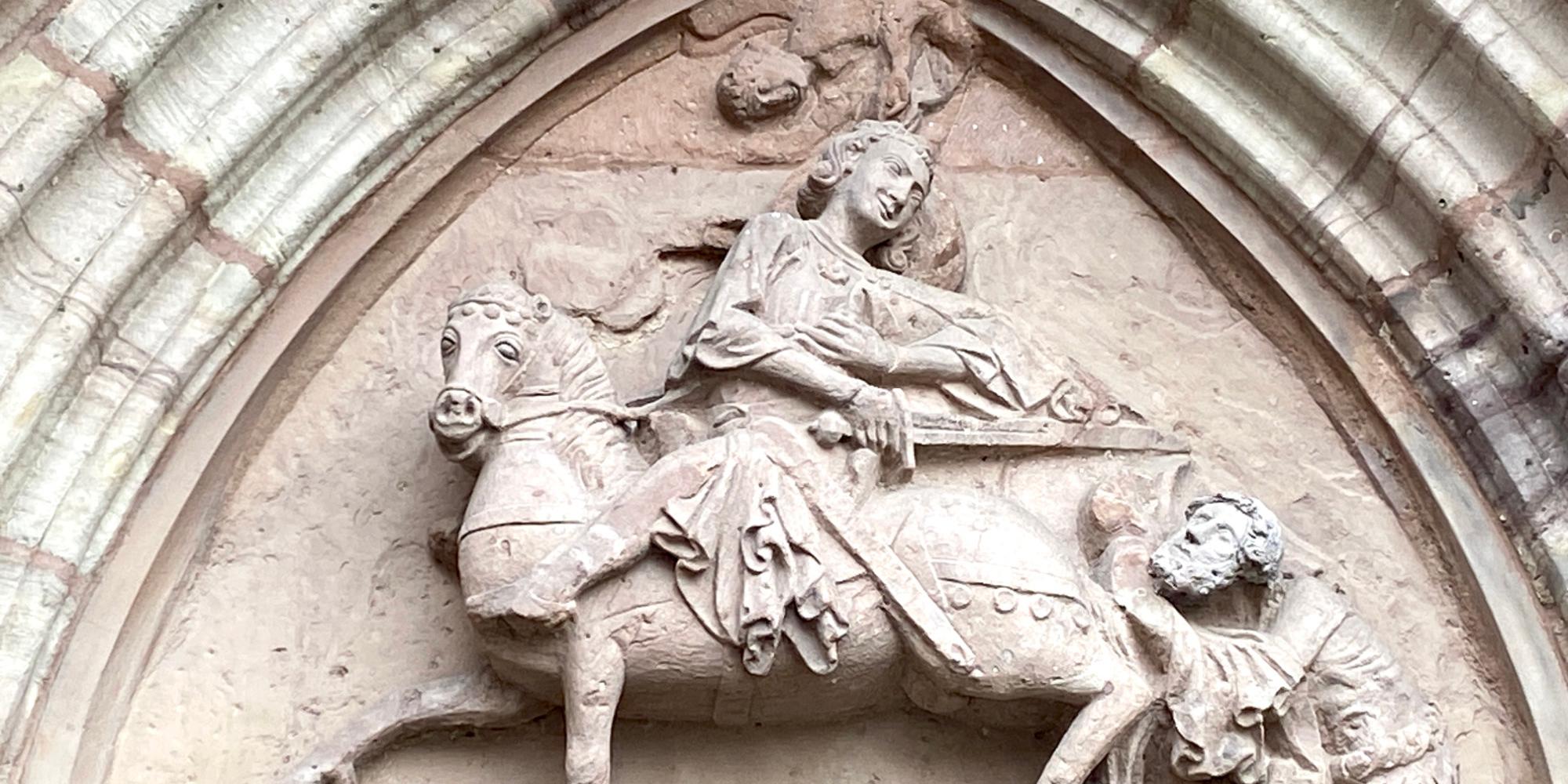 Das Foto zeigt ein steinernes Relief in einem spitzbogigen Rahmen, auf dem der heilige Martin auf einem Pferd sitzt und seinen Mantel mit einem frierenden Bettler teilt.