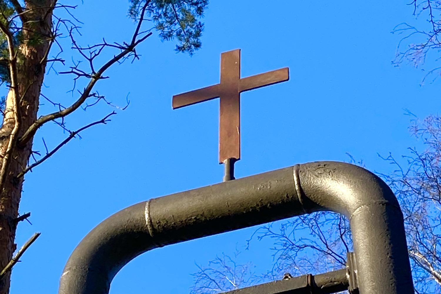 Kirchenglocke an einem Metallgestellt mit Kreuz, blauer Himmel und kahle Bäume im Hintergrund