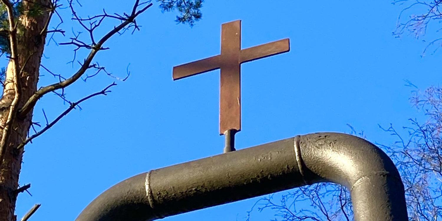 Kirchenglocke an einem Metallgestellt mit Kreuz, blauer Himmel und kahle Bäume im Hintergrund
