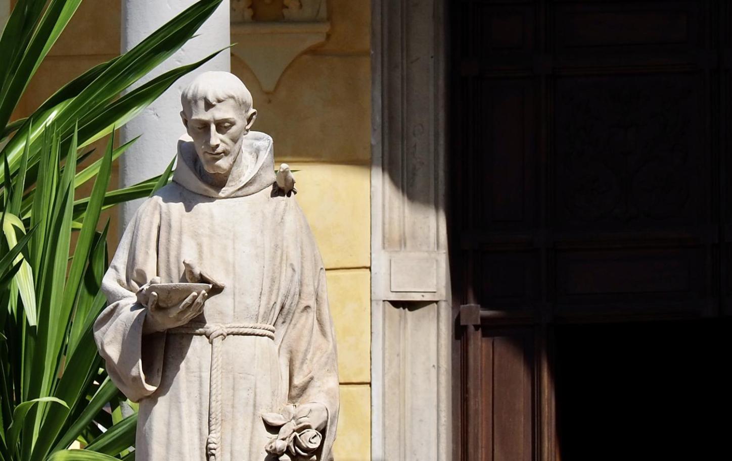 Steinstatue eines Mönchs mit Buch in sonnigem Hof neben einer Pflanze