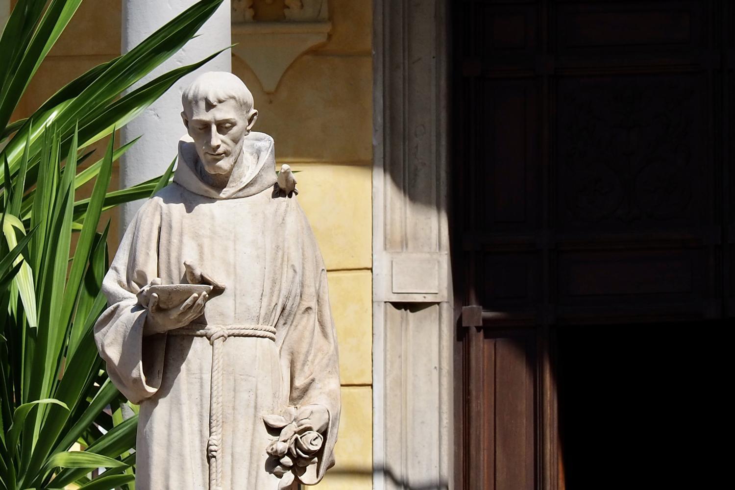 Steinstatue eines Mönchs mit Buch in sonnigem Hof neben einer Pflanze
