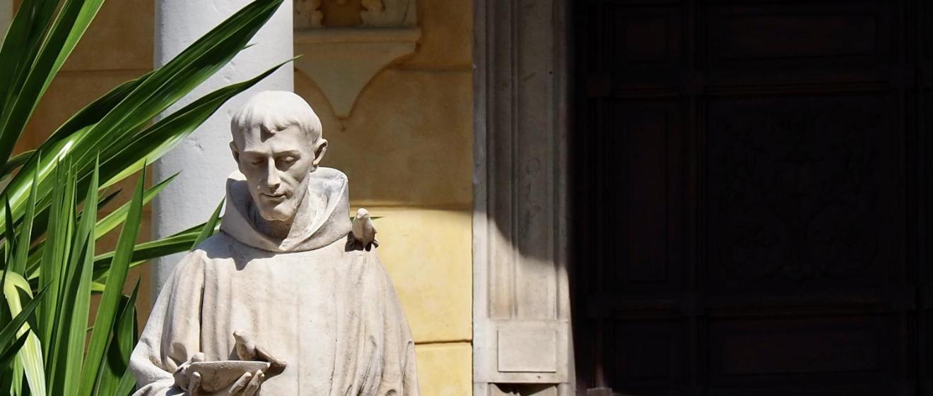 Steinstatue eines Mönchs mit Buch in sonnigem Hof neben einer Pflanze