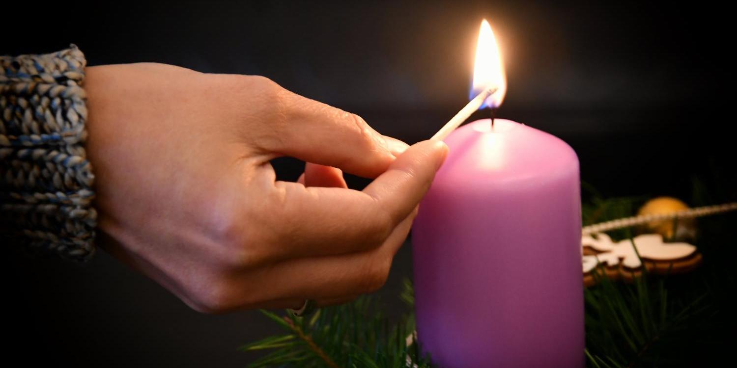 Das Foto zeigt eine Hand, die mit einem Streichholz eine große, lilafarbene Kerze anzündet, die in einem grünen Adventskranz mit goldenen Kugeln steht.
