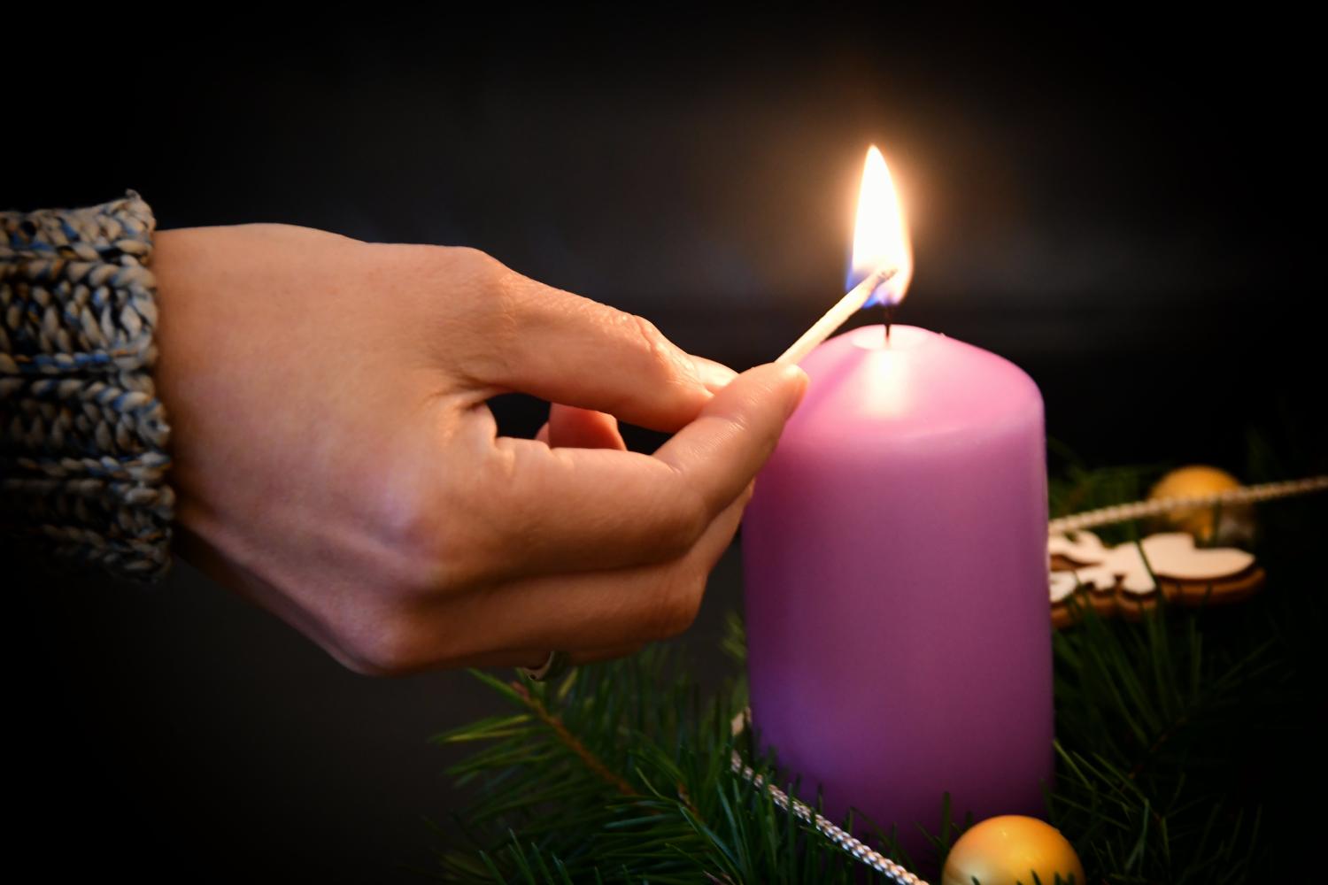 Das Foto zeigt eine Hand, die mit einem Streichholz eine große, lilafarbene Kerze anzündet, die in einem grünen Adventskranz mit goldenen Kugeln steht.