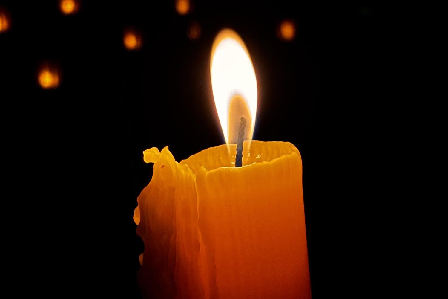 Eine einzelne, brennende Kerze leuchtet warm im Dunkel, umgeben von kleinen Lichtpunkten im Hintergrund.