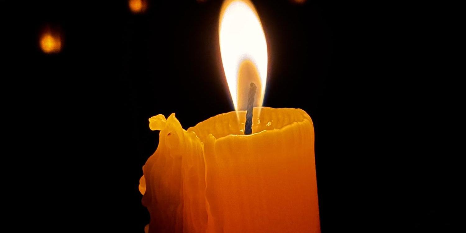 Eine einzelne, brennende Kerze leuchtet warm im Dunkel, umgeben von kleinen Lichtpunkten im Hintergrund.